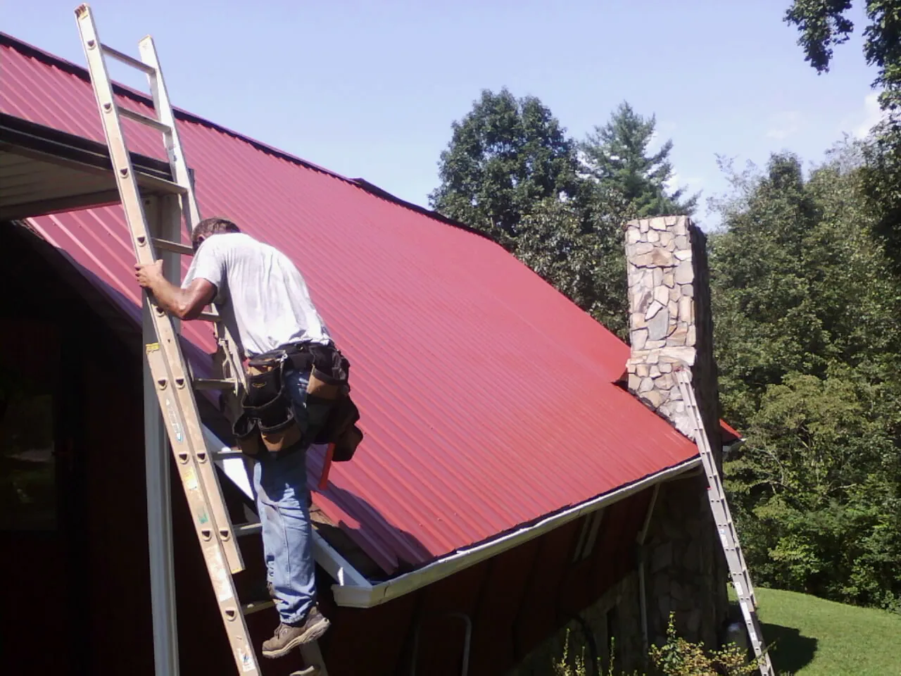 Roofer on a ladder beside a red metal roof and stone chimney — Soffit & Fascia Repair by Roofing Brothers in Topanga
